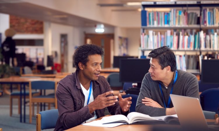 two male adult students talking at table in library