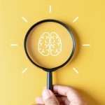 Hand holding magnifying glass against yellow background with brain illustration in centre of lens