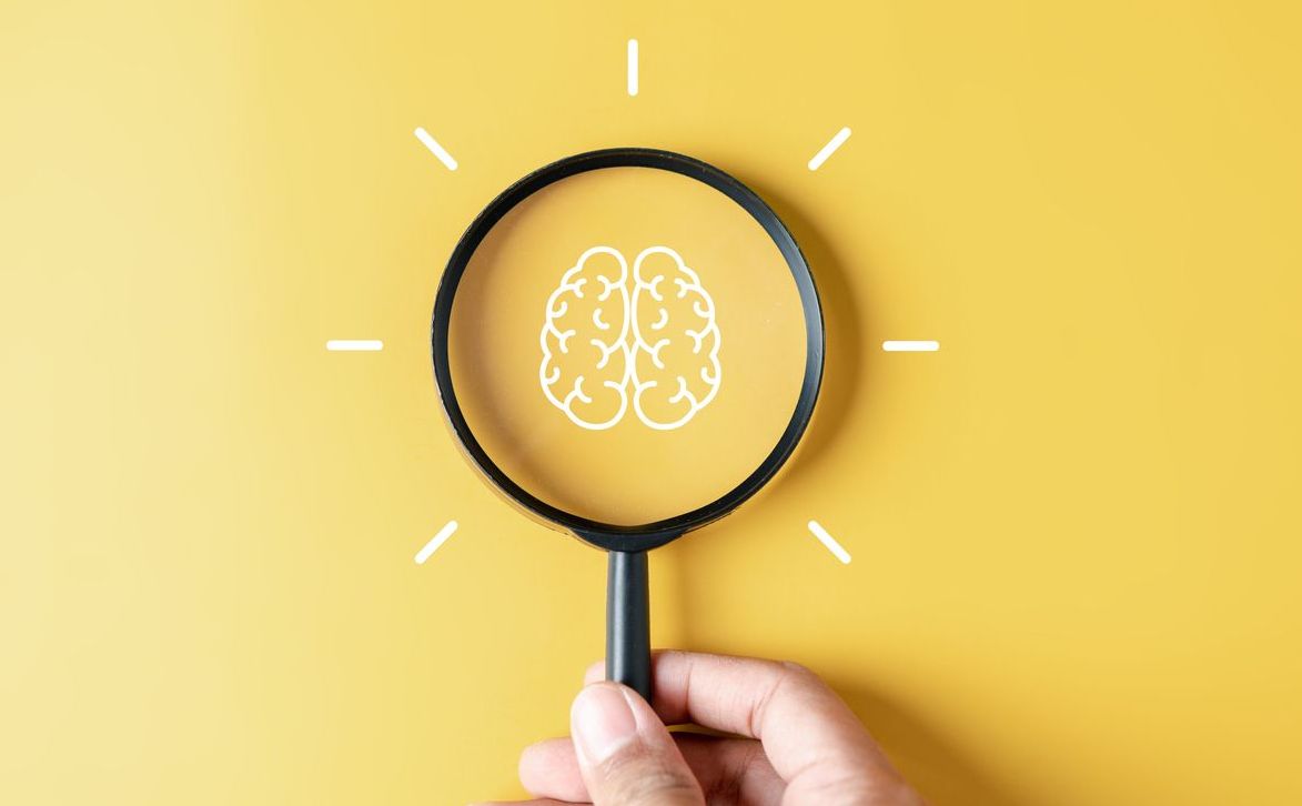 Hand holding magnifying glass against yellow background with brain illustration in centre of lens