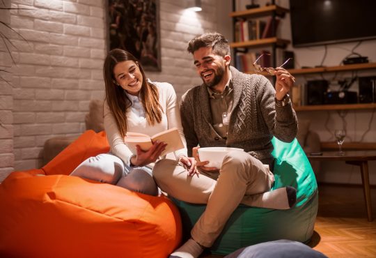 Happy couple smiling and reading book while sitting on lazy bags. Woman holding book. Living room interior.