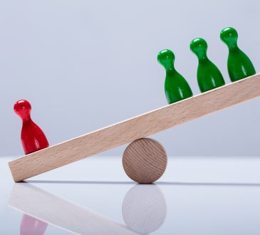 Red And Green Pawns Figures Balancing On Wooden Seesaw