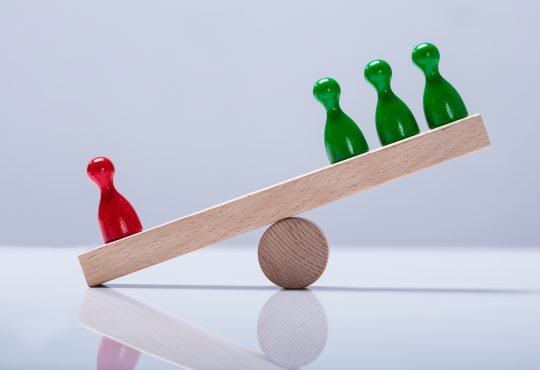 Red And Green Pawns Figures Balancing On Wooden Seesaw