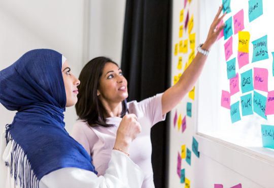 woman in hijab with woman not in hijab putting sticky notes on whiteboard