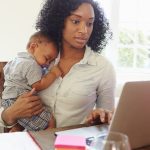 Mother With Baby Working In Office At Home Looking At Laptop