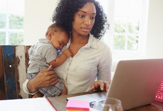 Mother With Baby Working In Office At Home Looking At Laptop