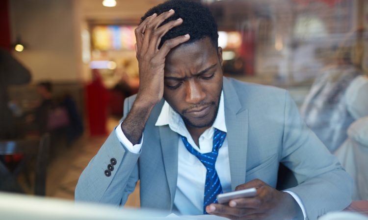 businessman looking stressed and resting his head on hand while typing message in smartphone sitting at counter in coffee shop,