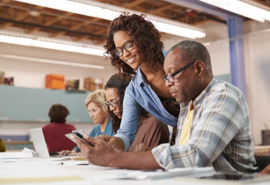 Teacher Helping Retired Senior Man Attending IT Class In Community Centre