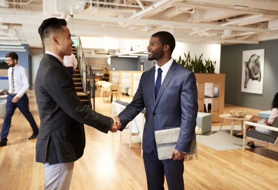 Two Businessmen Meeting And Shaking Hands In Modern Open Plan Office