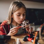 Girl wearing red hoodie looking circuit board throughout magnifying glass at home and building a robot