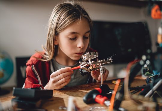 Girl wearing red hoodie looking circuit board throughout magnifying glass at home and building a robot