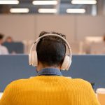 Rear view of busy brunette man in wireless headphones wearing bright sweater sitting at table in open space office a