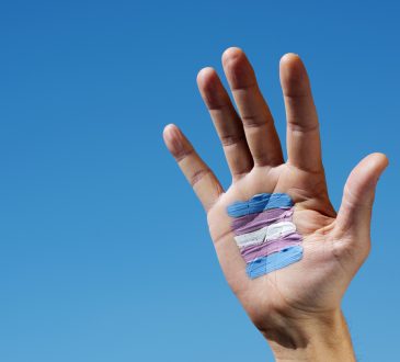closeup of a transgender flag painted in the palm of the hand of a young caucasian person, against the blue sky