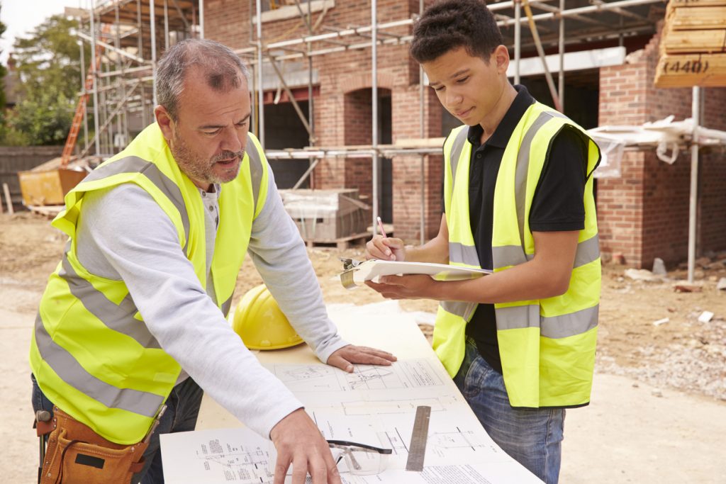 Builder On Building Site Discussing Work With Apprentice