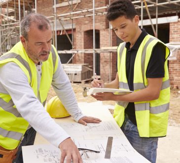 Builder On Building Site Discussing Work With Apprentice