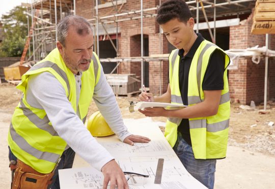 Builder On Building Site Discussing Work With Apprentice