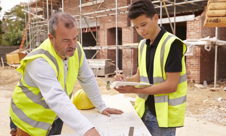 Builder On Building Site Discussing Work With Apprentice