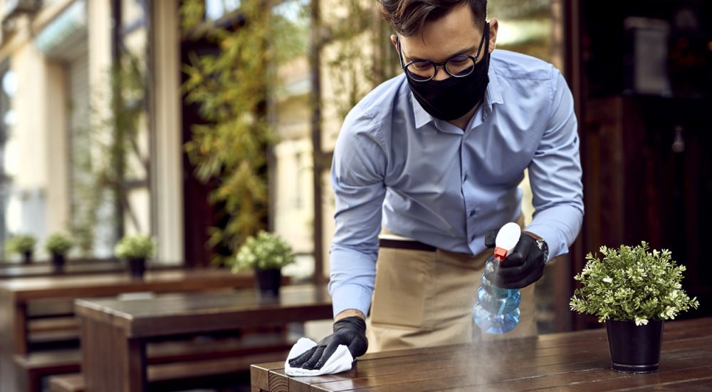 Waiter wearing protective face mask while disinfecting tables at outdoor cafe.
