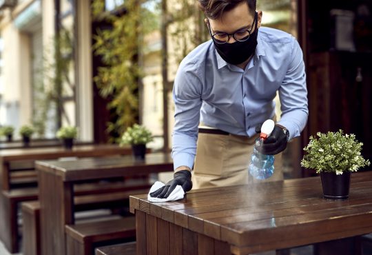 Waiter wearing protective face mask while disinfecting tables at outdoor cafe.