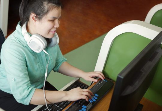 Woman with visual impairment wearing headphones using computer with refreshable braille display