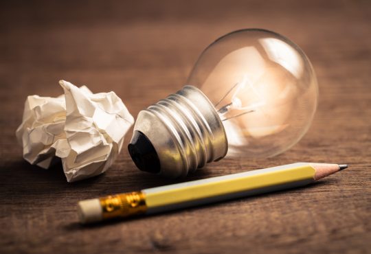 Pencil with glowing light bulb and crumpled paper on wood table