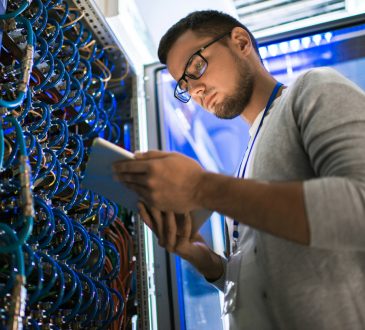 young man using digital tablet standing by server cabinet while working with supercomputer in blue light