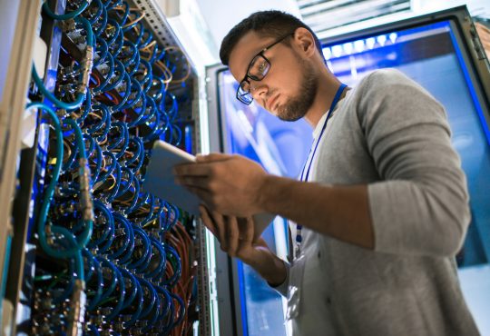 young man using digital tablet standing by server cabinet while working with supercomputer in blue light