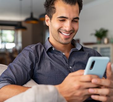 Cheerful businessman using smartphone while sitting on sofa at home.