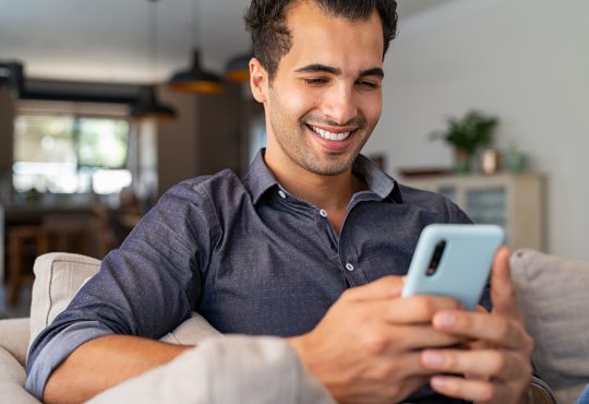 Cheerful businessman using smartphone while sitting on sofa at home.