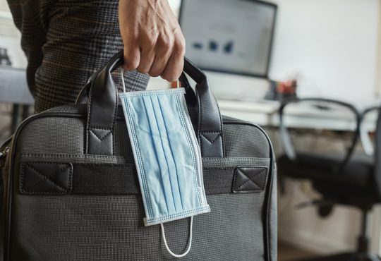 closeup of a young man in an office holding a briefcase and a surgical mask in his hand