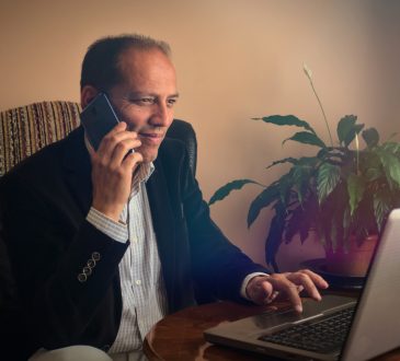 Happy and smiling senior man sitting on armchair phoning to a friend while browsing through internet with his laptop,