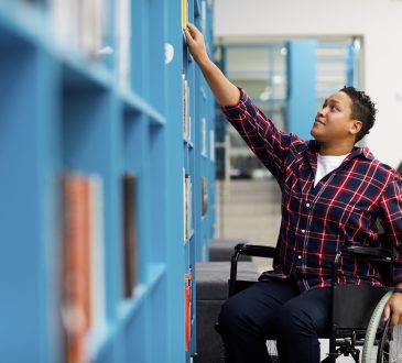 student in wheelchair choosing books while studying in college library