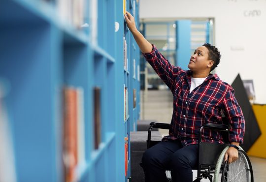 student in wheelchair choosing books while studying in college library