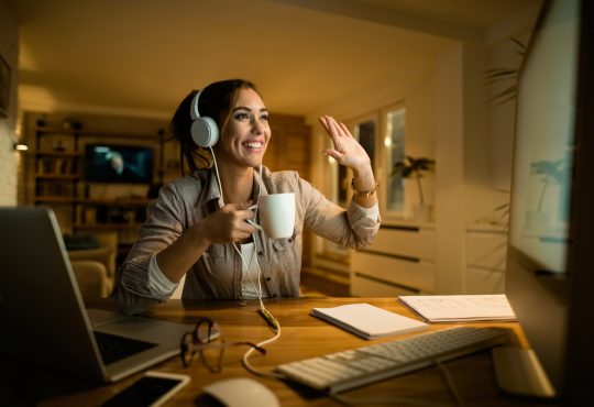 Happy woman drinking tea and waving to someone while having video call over desktop PC in the evening at home.