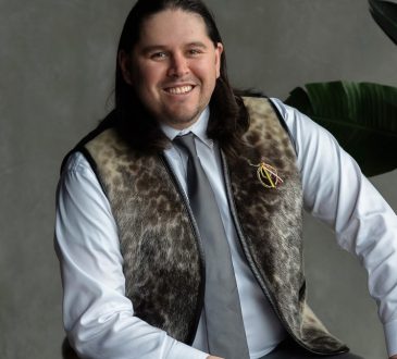 Michael Etherington sitting for a posed photo on grey background with green plant beside him