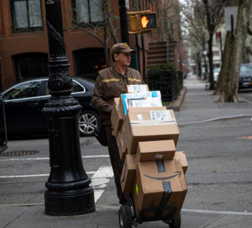 Middle aged delivery man in brown coveralls and hat, walking on the sidewalk of a small street with a trolly stacked with boxes for delivery
