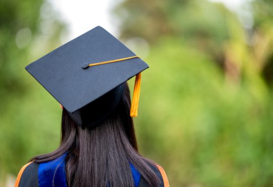 closeup behind a female college graduate wearing a black fringe gown and a black hat,
