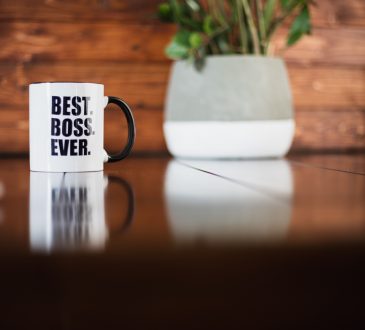 A coffee mug with Best Boss Ever printed on it on a table with plant, books and a laptop