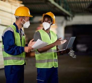 two men using laptop and communicating while working at lumberyard warehouse during COVID-19 pandemic.