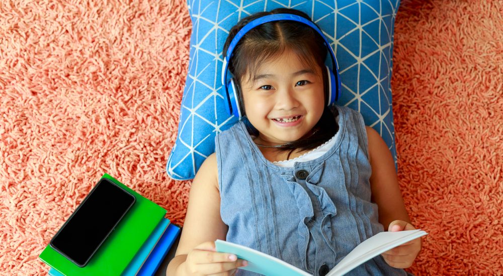 little girl lying on floor of bedroom reading and listening through headphones
