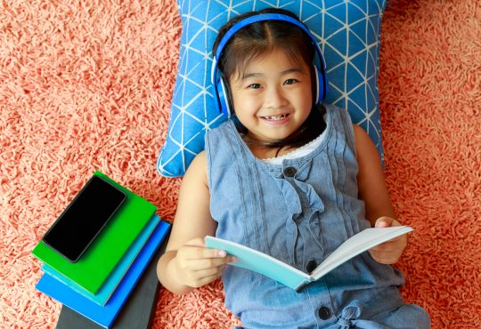 little girl lying on floor of bedroom reading and listening through headphones