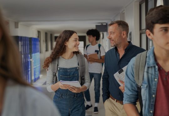 Female high school student and male teacher walking in hallway talking amid other students.