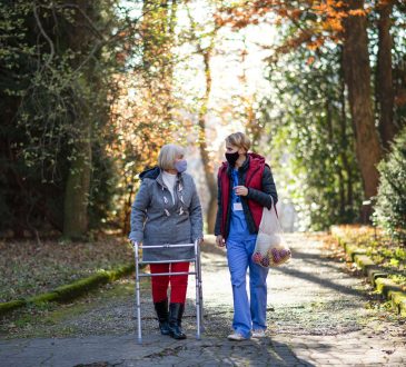 Senior woman using walker and female healthcare working walking outdoors on path in fall