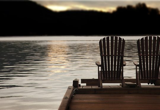 Adirondack chairs on a pier at sunset by the lake or ocean.