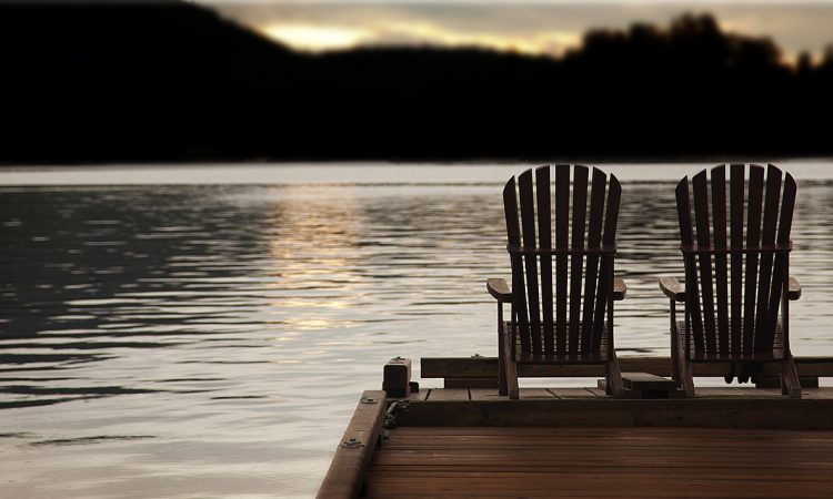 Adirondack chairs on a pier at sunset by the lake or ocean.