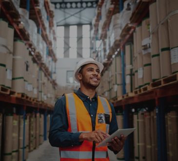Young man wearing safety jacket in warehouse holding clipboard.