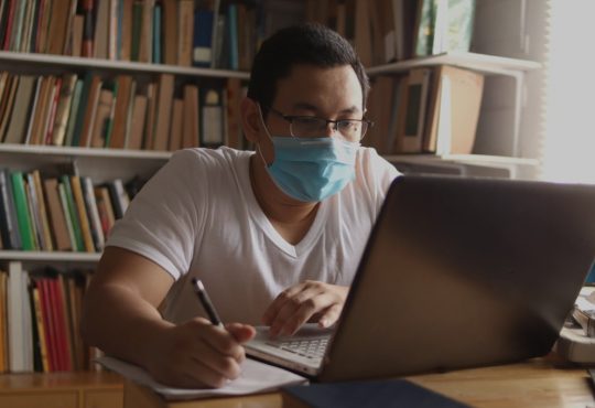 Man using laptop in library wearing mask.