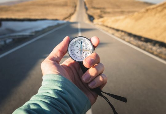 Compass in hand in front of mountain road background.