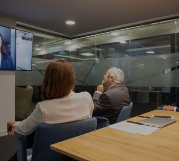People sitting in boardroom having conversation with woman on teleconferencing on tv screen.