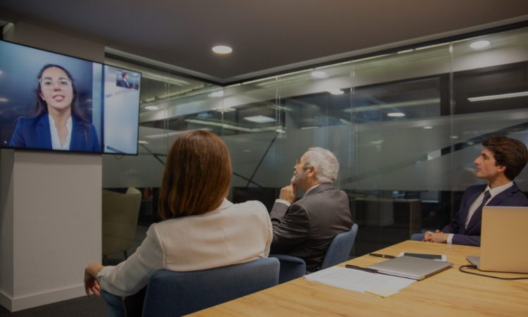 People sitting in boardroom having conversation with woman on teleconferencing on tv screen.