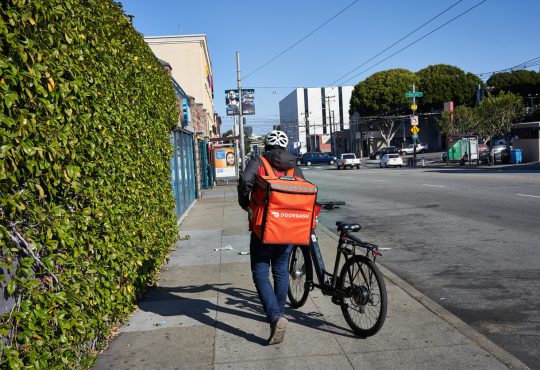 A DoorDash delivery worker walks his bike along the road.